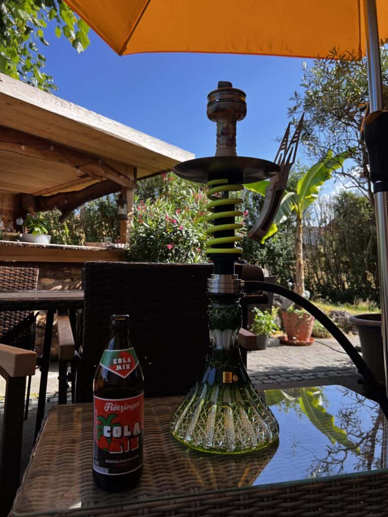 Rauchsäule mit Wasserpfeife auf einem Tisch im Garten unter einem Sonnenschirm.