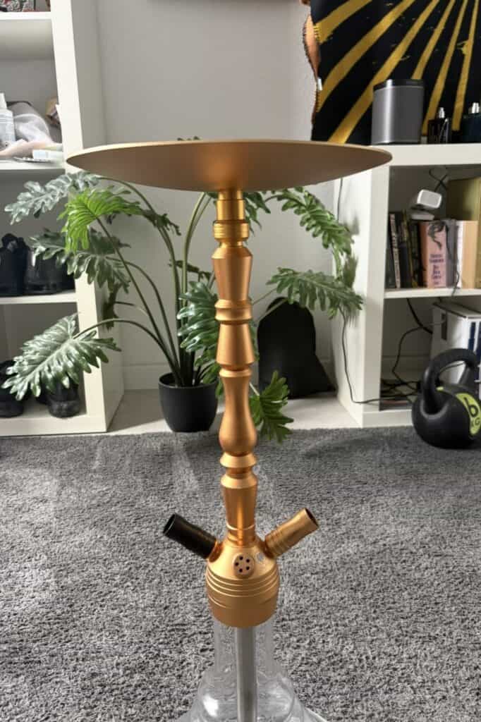 Goldene Shisha mit Ablagefläche auf Teppichboden, Hintergrund mit Pflanzen und Bücher.
