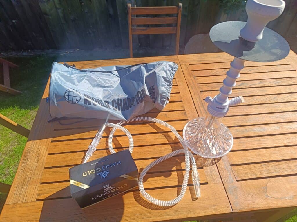 Shisha mit Rauchsäule und Zubehör auf Holztisch im Garten.