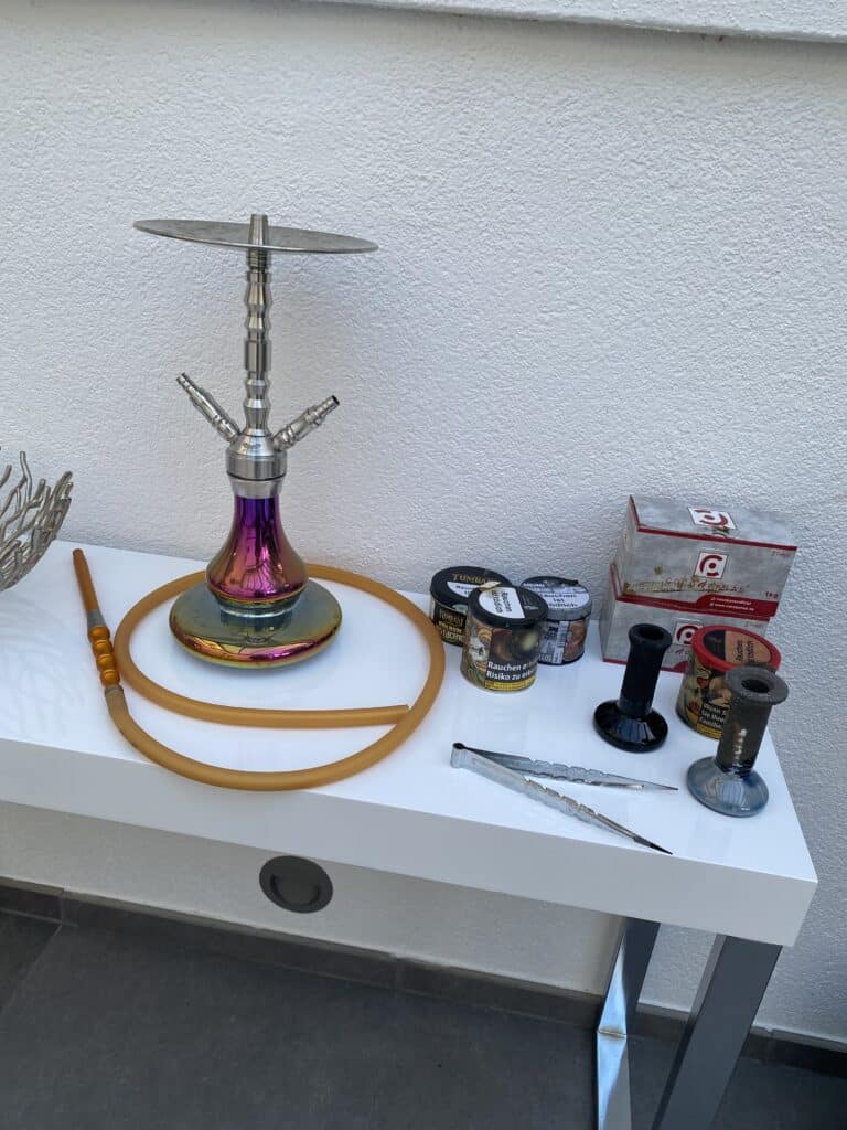 Shisha mit Zubehör und Tabak auf weißem Tisch, inklusive Kohle, Tabak und Ersatzteile.