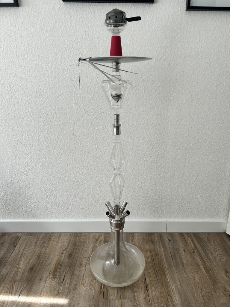 Wasserpfeife mit Glas- und Metallkomponenten, hoher Stand, auf Holzboden vor weißer Wand.