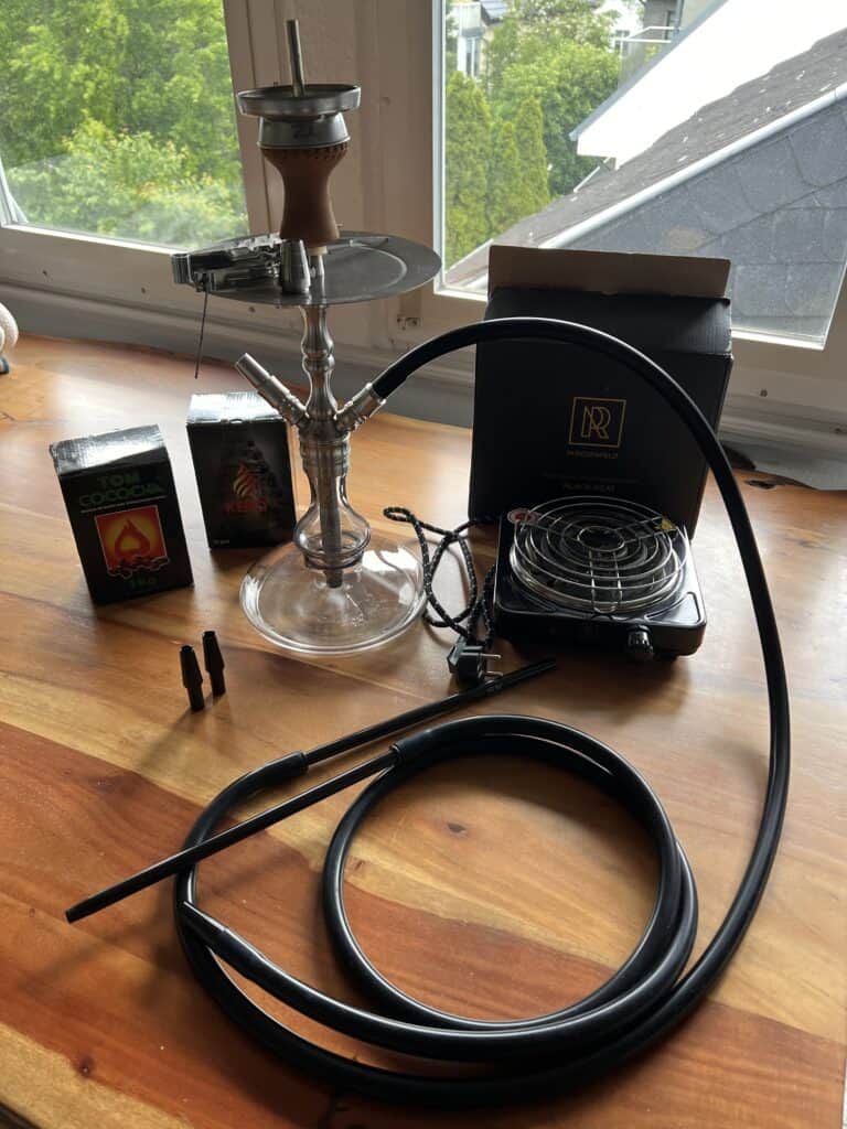 Shisha mit Rauchsäule, Schlauch und Zubehör auf Holztisch vor Fenster.
