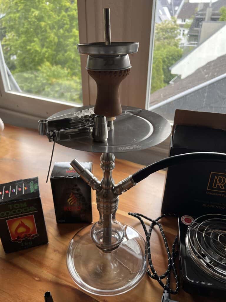 Rauchende Shisha mit Glasbowl, Schlauch und Tabakbox auf Fensterbank.