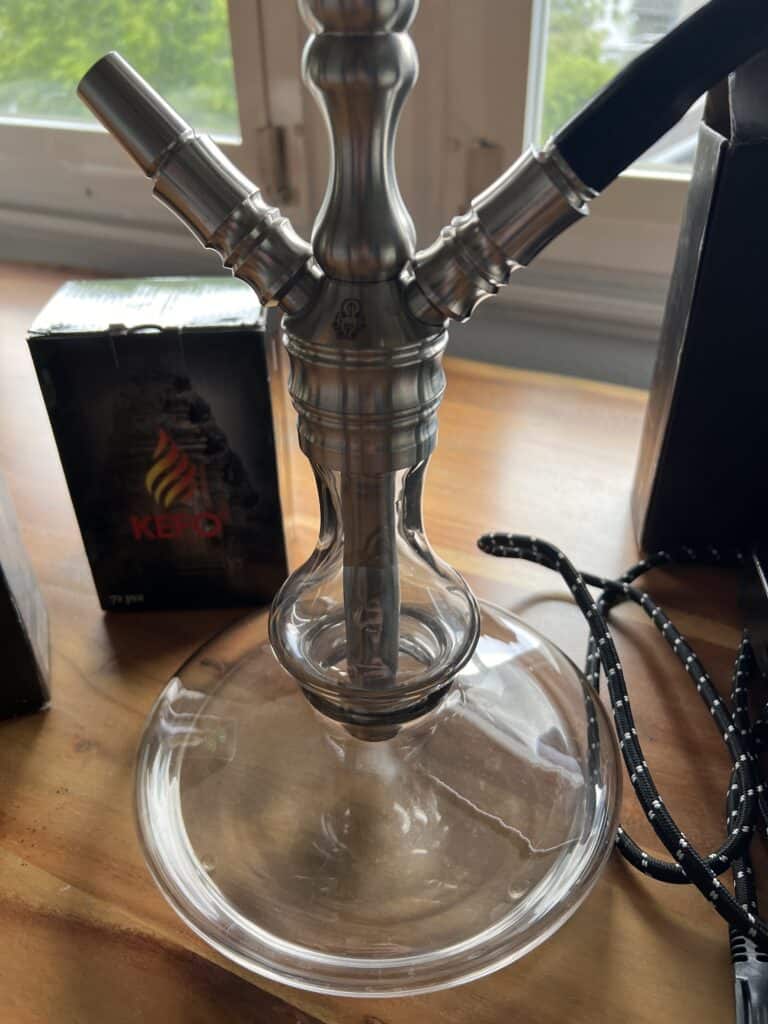 Moderne Shisha mit Metall- und Glasdesign auf Holztisch, Blick aus Fenster im Hintergrund.