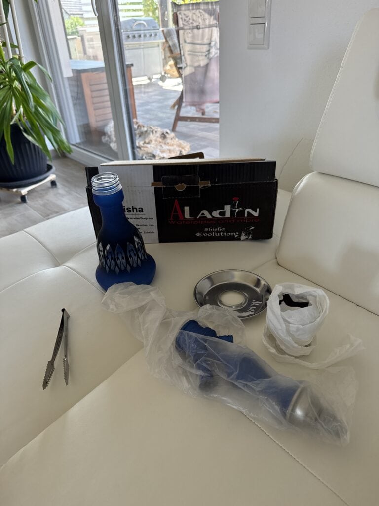 Neue Shisha mit Glas, Kohlezange, Tabak und Zubehör auf weißem Sofa.