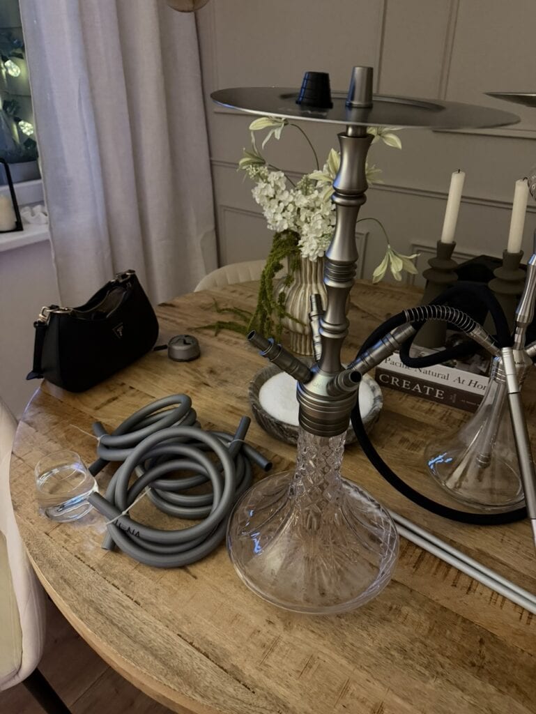 Shisha-Kohlenzange und Glasbowl auf Holztisch, Dekoration im Hintergrund.