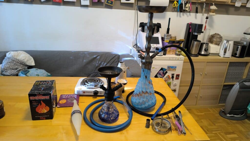 Bunte Shisha mit blauer Glasbowl und Zubehör auf Tisch.