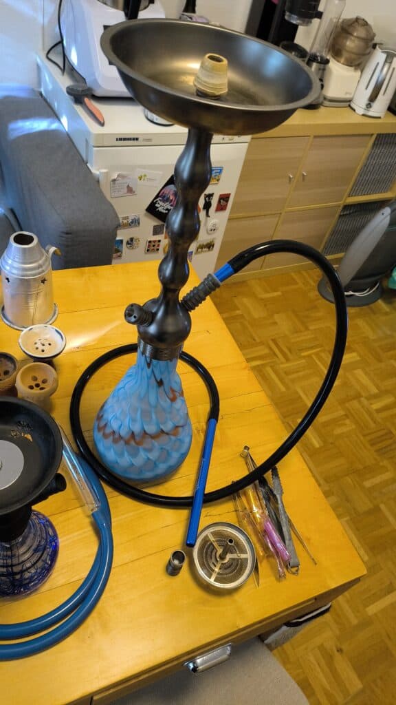 Shisha mit blauer Glasbowl, Schlauch und Zubehör auf Tisch.