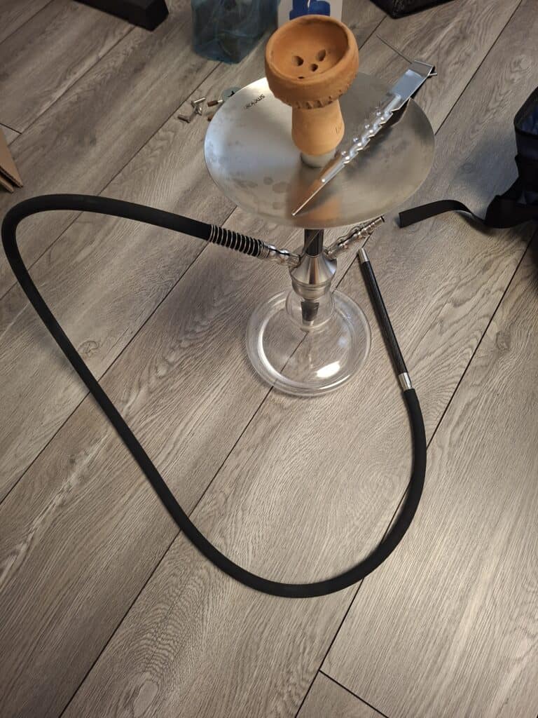 Shisha mit Glasbowl, Schlauch und Zange auf Holzboden.