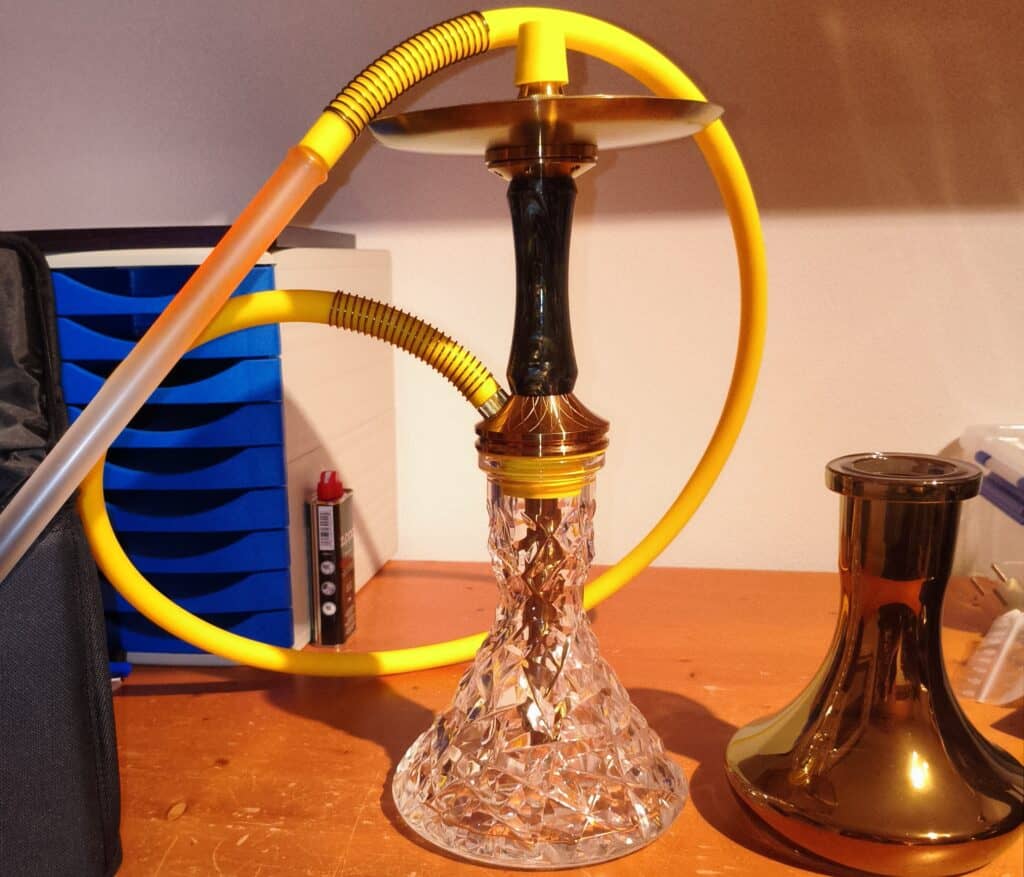 Shisha mit Glasbowl, Schlauch und Zubehör auf Tisch.