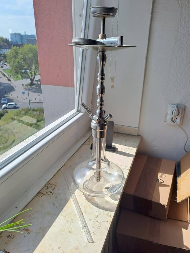 Shisha mit Glasbowl auf Fensterbank, sonniger Tag, Stadtblick im Hintergrund.