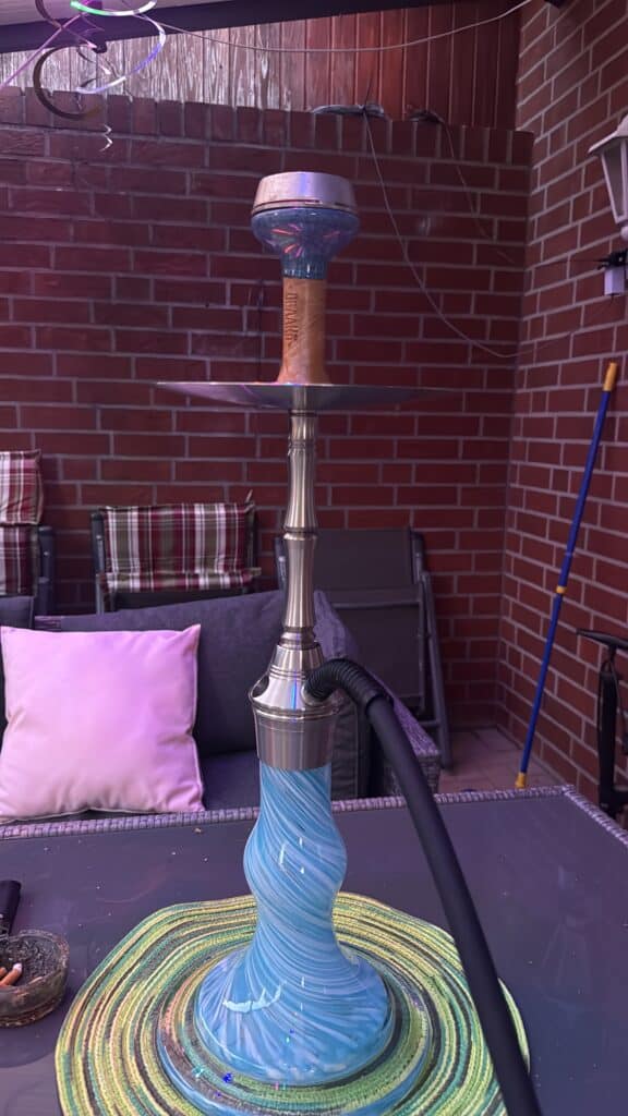 Shisha-Kopf mit bunter Glasbowl auf Terrasse, Backsteinwand im Hintergrund.