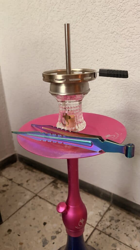 Shisha-Glasbowl mit Metallring und Zubehör auf pinkem Tisch.