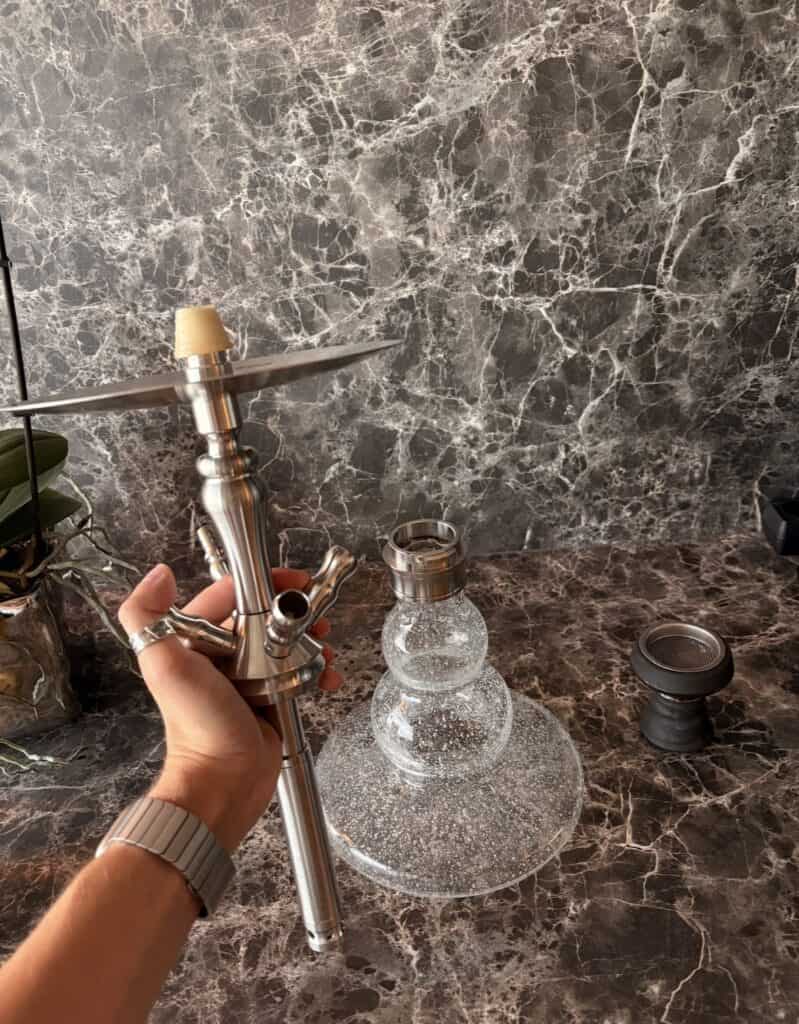 Hand hält Shisha mit Metallrohr und Glasbowl auf Marmoroberfläche.