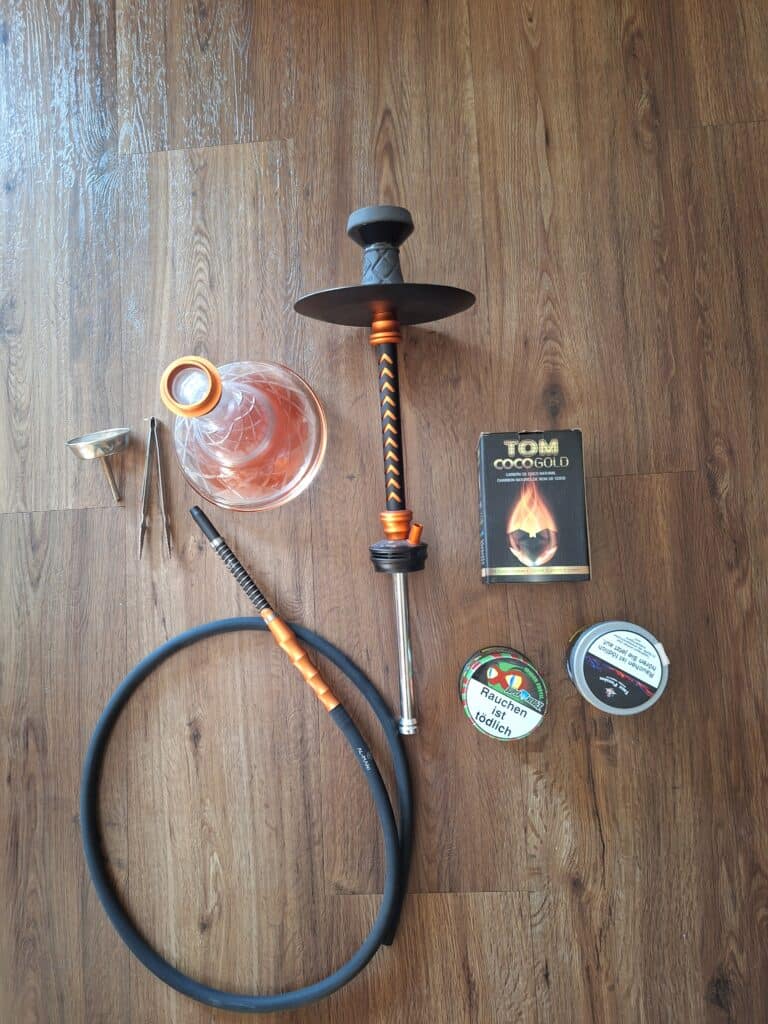 Shisha mit Glasbowl, Schlauch, Dichtung und Tabakpackung auf Holztisch.