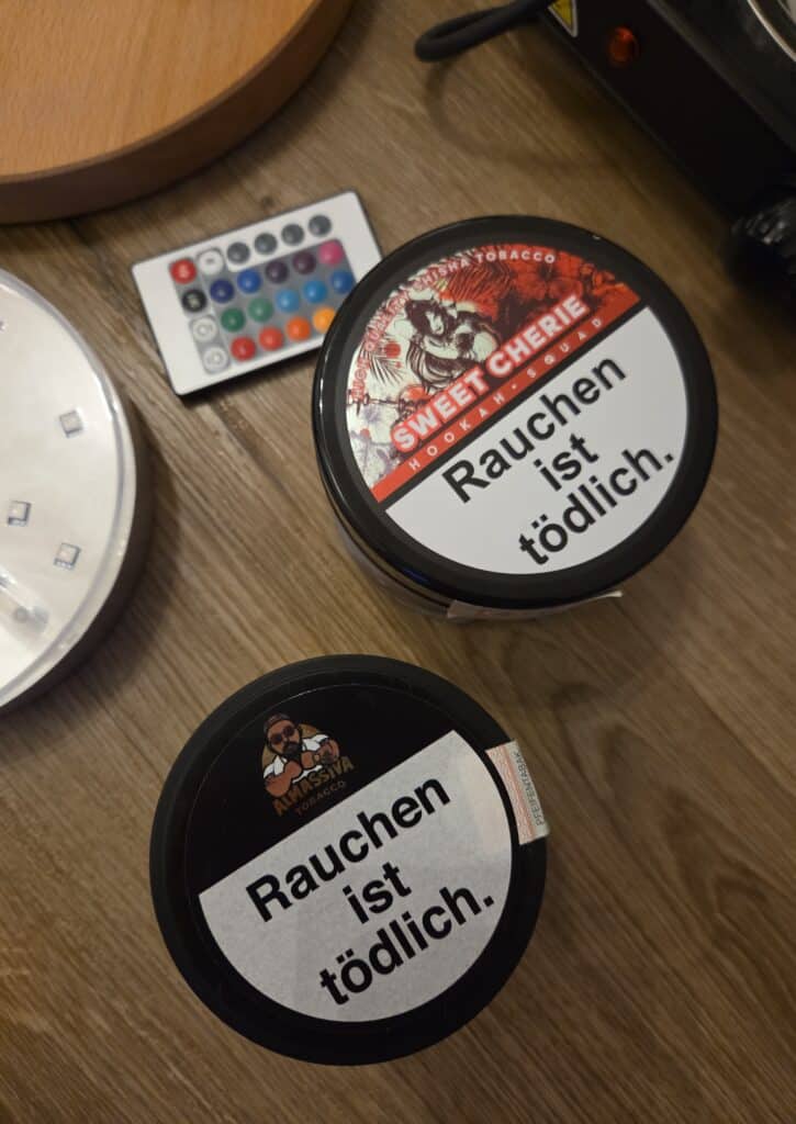 Zwei Shisha-Glasbowl mit Warnhinweisen und Farbpalette auf Holzoberfläche.