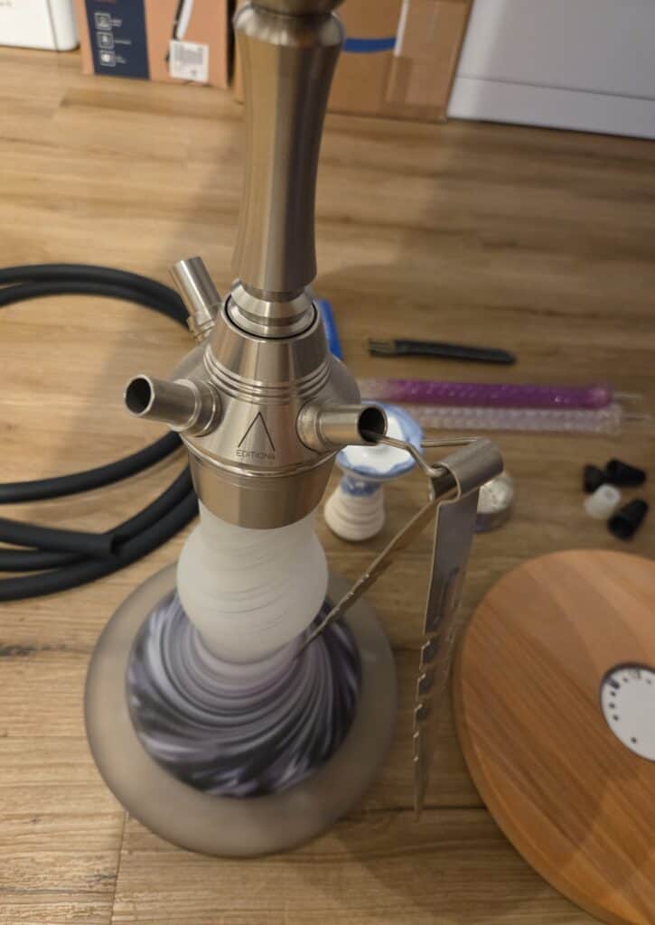 Shisha mit Metallkopf, Glasbowl und Zubehör auf Holzoberfläche.