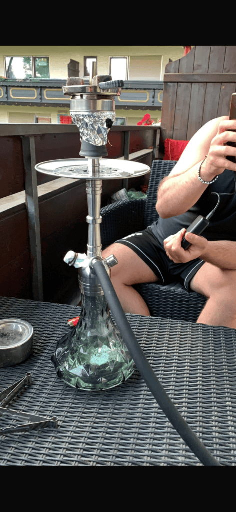 Shisha mit Glasbowl auf einem Tisch im Garten, Person hält den Schlauch.