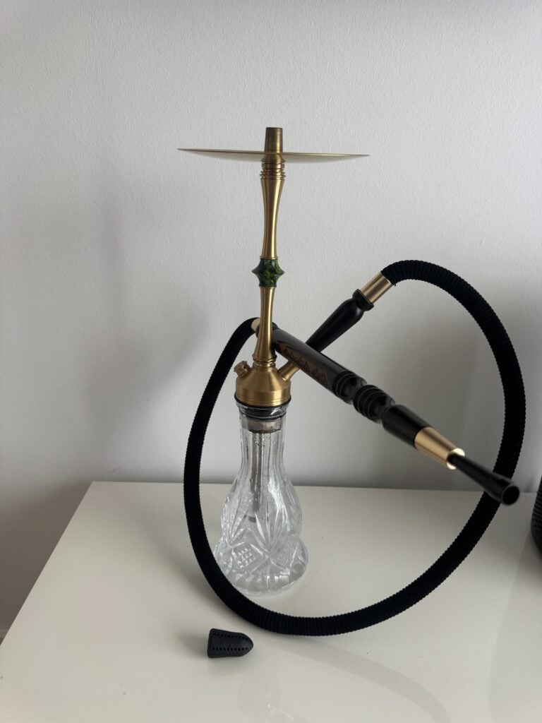 Shisha mit Glasbowl, Schlauch und Metallhaken auf weißem Tisch.