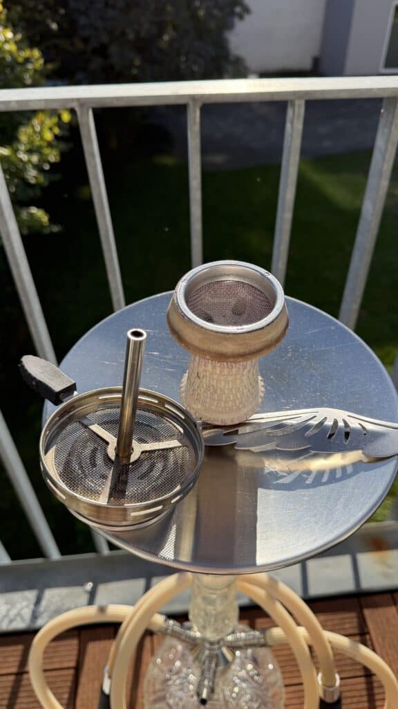 Shisha mit Glasbowl, Metallzubehör und Tabak auf Balkon.