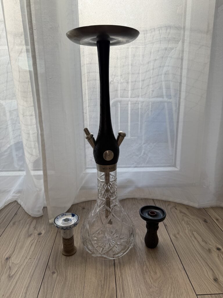 Shisha-Kopf mit Glasbowl und Zubehör, auf Holzboden vor Fenster.