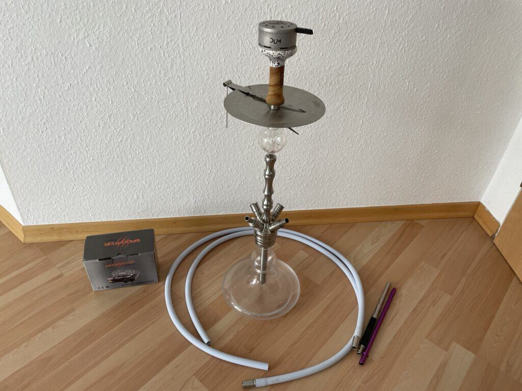 Shisha mit Glasbowl, Schlauch und Zubehör auf Boden.