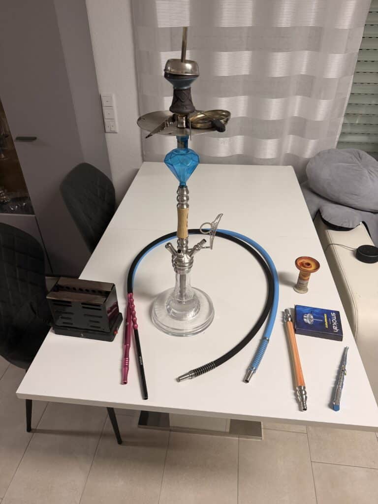 Shisha mit Glasbowl, Schlauch und Zubehör auf Tisch.