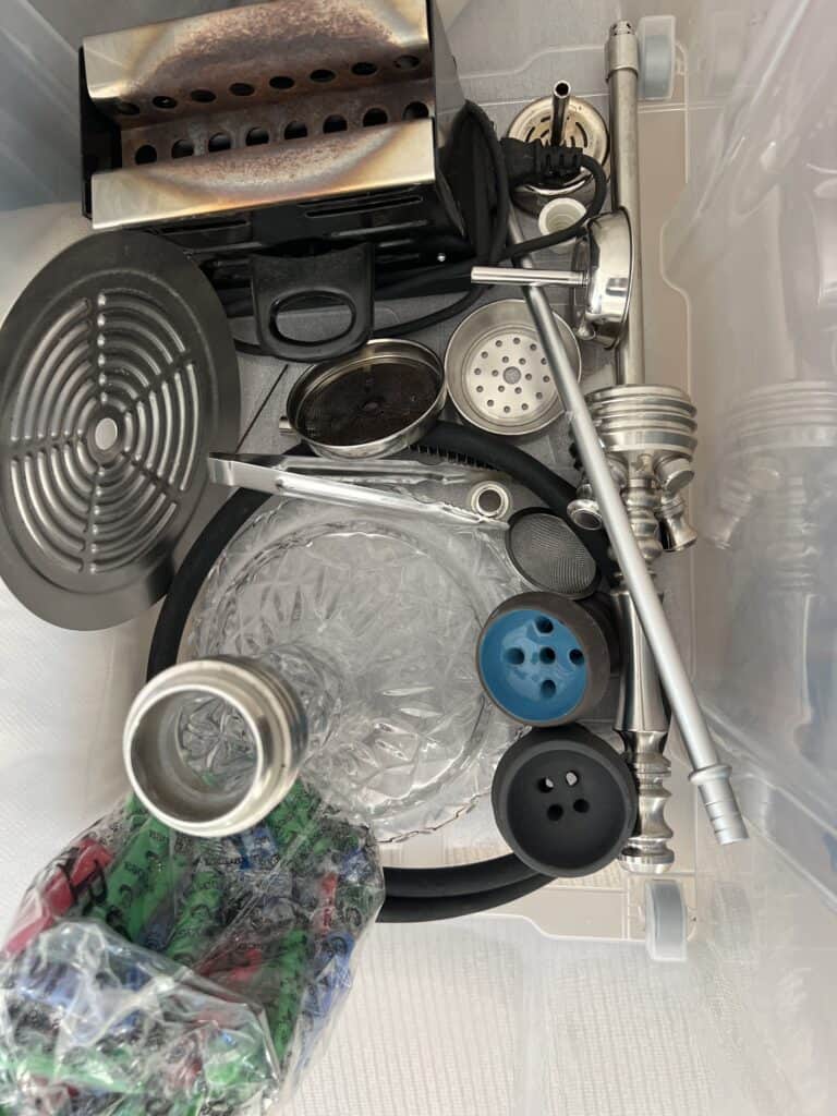Verschiedene Shisha-Zubehörteile in einer Box, inklusive Glasbowl, Schlauch und Kohlezange.