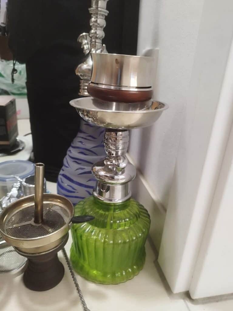 Shisha-Glasbowl mit Metallaufsatz und Zubehör auf Tisch.