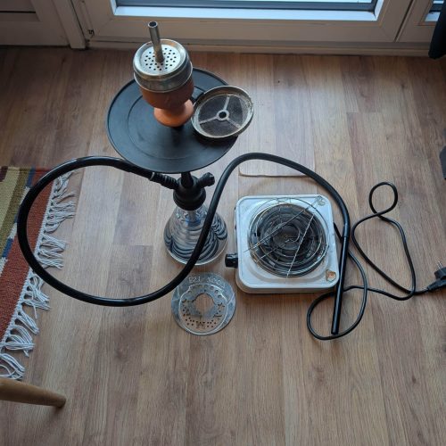Rauch-Hookah mit Metall- und Glaszubehör, Steh- und Schlauchteile, auf Holzboden vor Fenster.