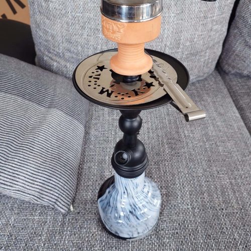 Alte Wasserpfeife mit Metallkopf und Schlauch, auf Couch sitzend.