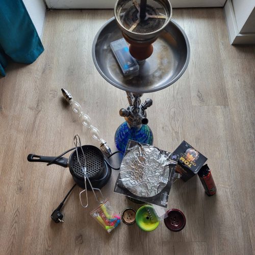 Shisha mit Wasserpfeife und Zubehör, bereit zum Rauchen, auf Holzboden vor Fenster.