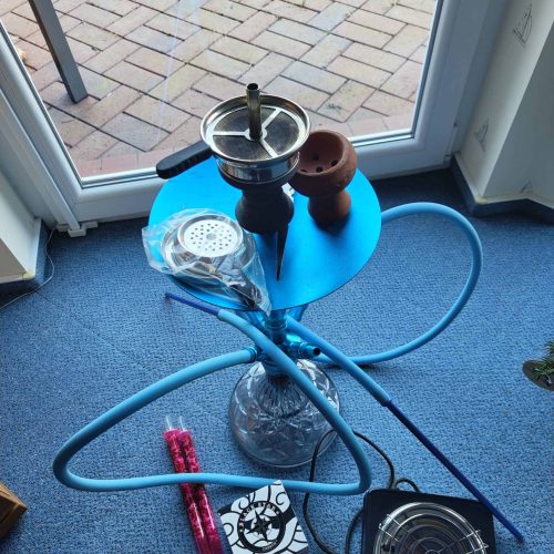 Shisha-Set mit Kohle, Tabak und Zubehör auf Teppich vor Fenster.