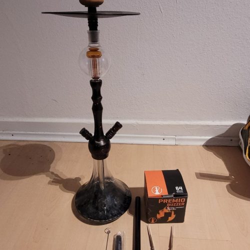 Shisha mit Zubehör, inklusive Kohle und Zange, auf Boden stehend.