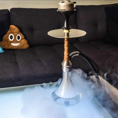 Shisha mit Rauchentwicklung auf einem Tisch, gemütliches Wohnzimmer mit schwarzem Sofa im Hintergrun.