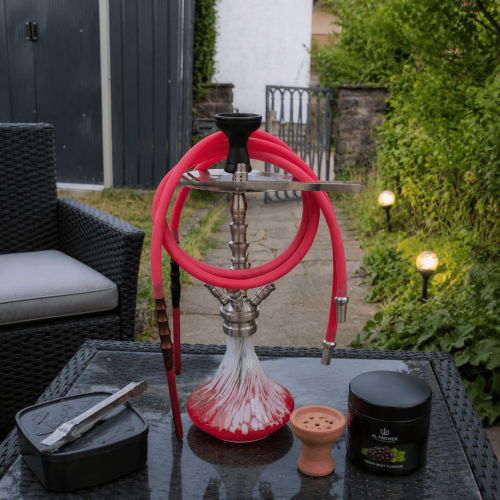 Shisha mit roter Glasbowl auf Tisch im Garten, Zubehör sichtbar.
