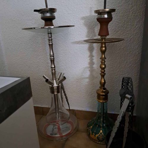 Zwei Shisha-Kohlenzangen neben einer Shisha, eine aus Edelstahl, die andere aus Messing.