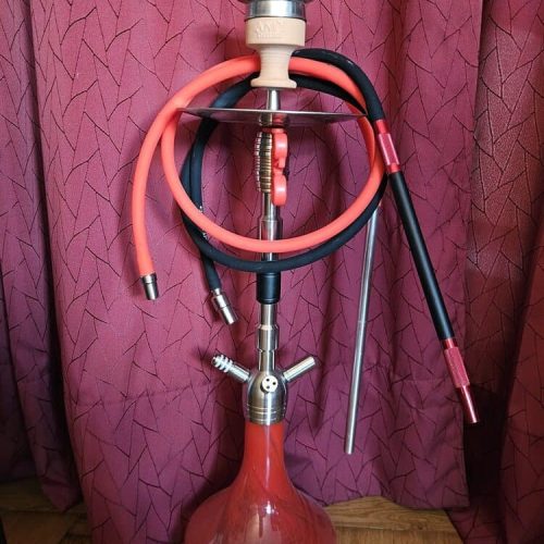 Shisha mit mehreren Schläuchen und Glasgefäß, auf Holzboden vor pinkem Vorhang.