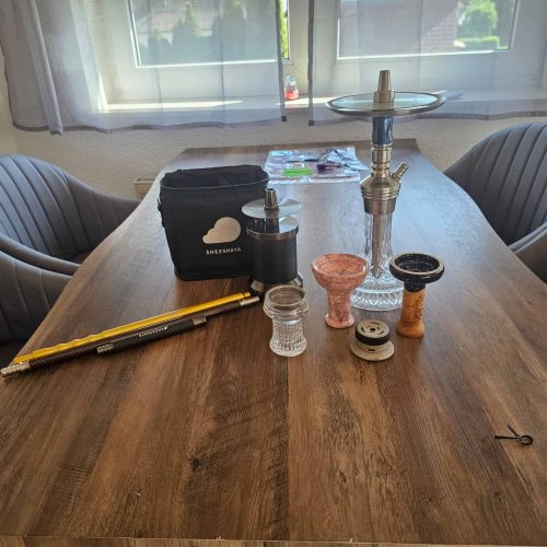 Rauchsäule mit Shisha-Set und Zubehör auf Holztisch, Fenster im Hintergrund.