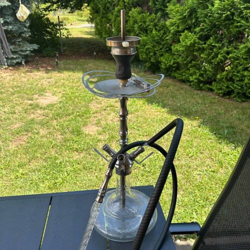 Shisha mit Glasbowl auf Tisch im Garten, grüne Bäume im Hintergrund.