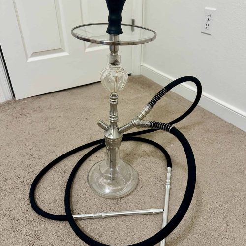 Wasserpfeife mit Glasbowl, Metallstamm und Schlauch, auf Teppichboden im Raum.