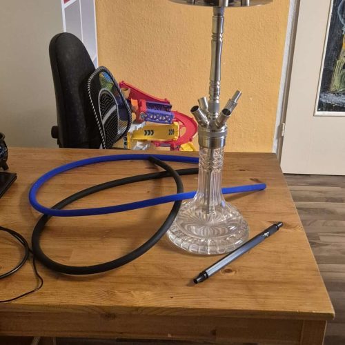 Wasserpfeife mit Glasaufsatz auf Holztisch, Zubehör und Dartboard im Hintergrund.
