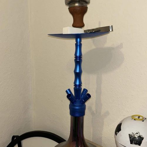Rauchende Shisha mit blauer Rauchsäule und Kopf, auf einem Tisch.