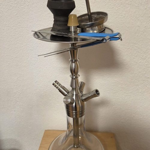 Shisha mit Glasbowl, Metallstücken und Zubehör auf Tisch.