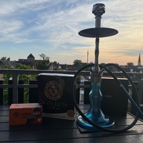 Shisha mit Glasbowl, Kohle und Zubehör auf Balkon bei Sonnenuntergang.