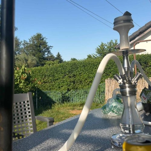 Rauchende Wasserpfeife auf einem Gartentisch unter Sonnenschirm.