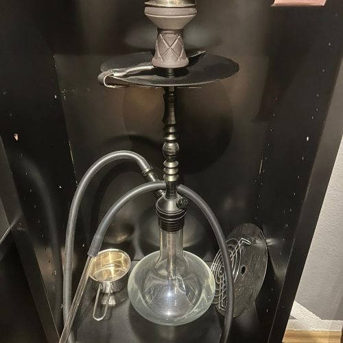 Shisha mit Glasballon, Schlauch und Tabakzubehör auf schwarzem Regal.