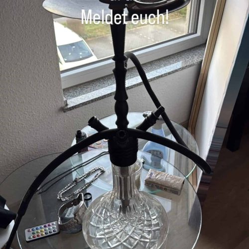 Schwarzer Shisha-Kopf mit Glasbasis, auf Fensterbank in Innenraum platziert, mit Zubehör im Hintergr.