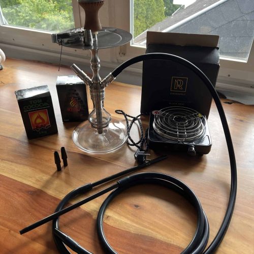 Shisha mit Rauchsäule, Schlauch und Zubehör auf Holztisch vor Fenster.