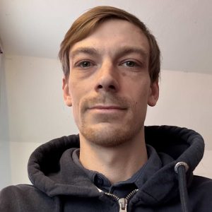 Junger Mann trägt dunklen Hoodie, Blick in die Kamera, kurze Haare.